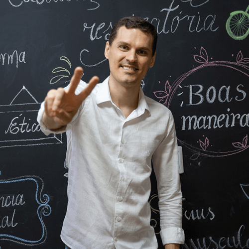 Homem de pele clara, camisa branca de manga longa, cabelo curto fazendo sinal de V com a mão direita